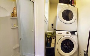 Un área de lavandería con dos lavadoras apiladas, una puerta y repisas con artículos de aseo.