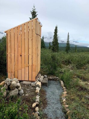 A wooden outhouse with a sloped roof, surrounded by a gravel path and a rock border, set in a natural landscape with trees and mountains in the background.