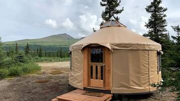 En cirkulær mongolsk tipi med en trædør og en rampe, omgivet af et naturområde med træer og bakker.