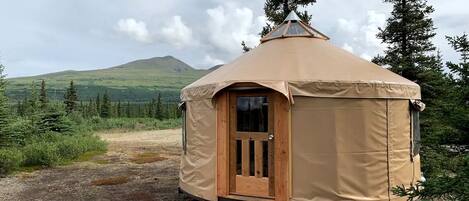 A circular yurt with a wooden door and a ramp, surrounded by a natural landscape with trees and hills.