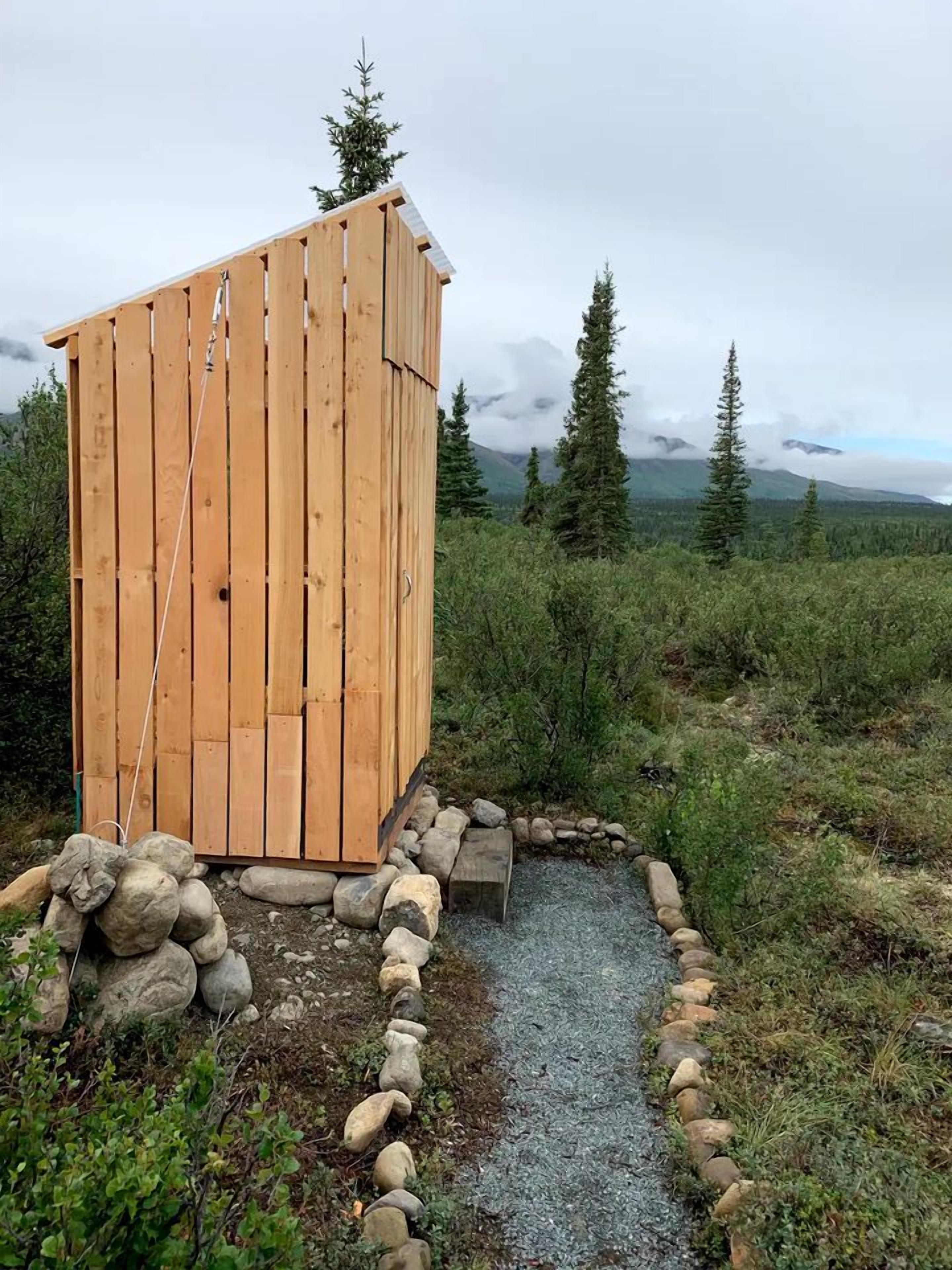 Et træ-toilet med skråt tag, omgivet af en grussti og en kant af sten, beliggende i et naturområde med træer og bjerge i baggrunden.
