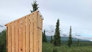 Puinen ulkohuussi, jossa on kalteva katto, ja jonka ympärillä on soraa ja kiviä reunuksena. Se sijaitsee luonnonmaisemassa, jonka taustalla näkyy puita ja vuoria.