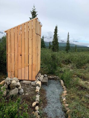 Un letrina de madera con techo inclinado, rodeada por un camino de grava y un borde de piedras, situada en un paisaje natural con árboles y montañas al fondo.
