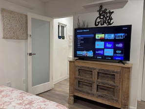 A television on a wooden stand in a room with a door, a wall with a decorative sign, and a floral bedspread. - Poolside Guesthouse in “The Valley” of LA – Cozy & Quiet Retreat (Los Angeles)