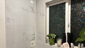 Een keuken met marmeren tegels, een raam met uitzicht op een bladerpatroon, een groene plant en keukengerei.