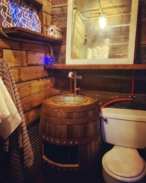 A rustic bathroom with a wooden barrel sink, a toilet, and a mirror above the sink.