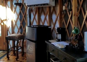 A compact kitchen with a refrigerator, a wooden stool, and a small table with kitchenware.