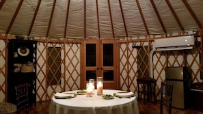 A round table with candles, plates, and cutlery in a tent with wooden lattice walls and a central door.