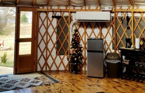 A cozy interior with a wooden floor, a refrigerator, a small Christmas tree, and a door leading outside. - Luxurious Lakefront Yurt Rental for a Romantic Getaway near Grapevine, Texas (Southlake)