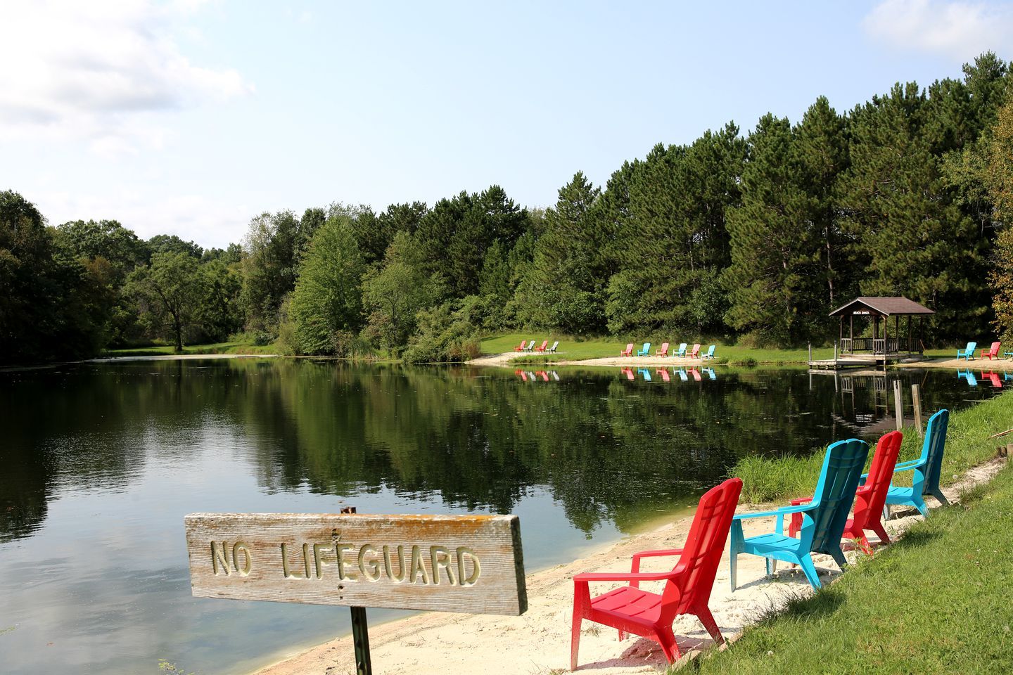 Uitzicht op een meer met een bord 'GEEN BADMEESTER', een steiger en kleurrijke Adirondack-stoelen.