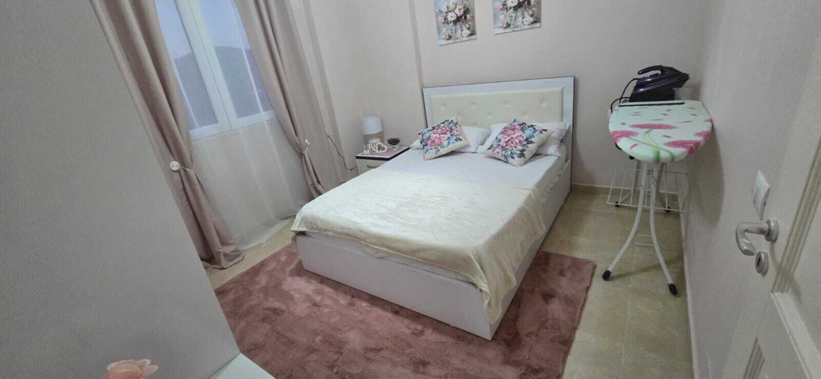 Quarto com cama, mesa de cabeceira, luminária, tábua de passar roupa na parede e janela com cortinas.