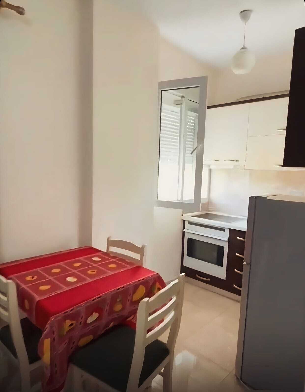 A small kitchen with white cabinets, a white oven, and a refrigerator. There is a dining table with a red tablecloth and white chairs.