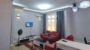 A modern living room with a flat-screen TV, a red sofa, a coffee table, and a dining area with a table and chairs. - Legacy Homes 1B Apartment (Dar es Salaam)