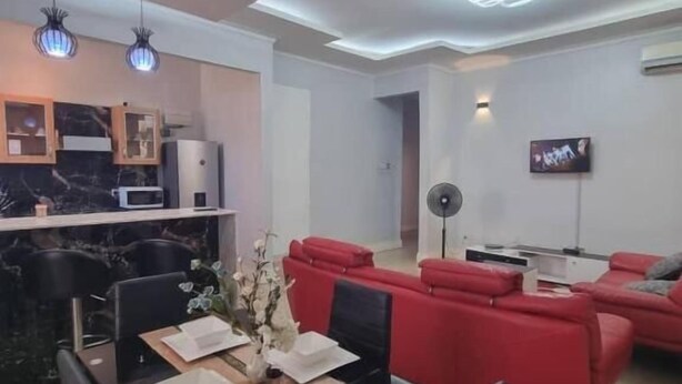 A modern living room with a dining area, red sofas, a TV, and a kitchen with marble countertops. - Legacy Homes 1B Apartment (Dar es Salaam)