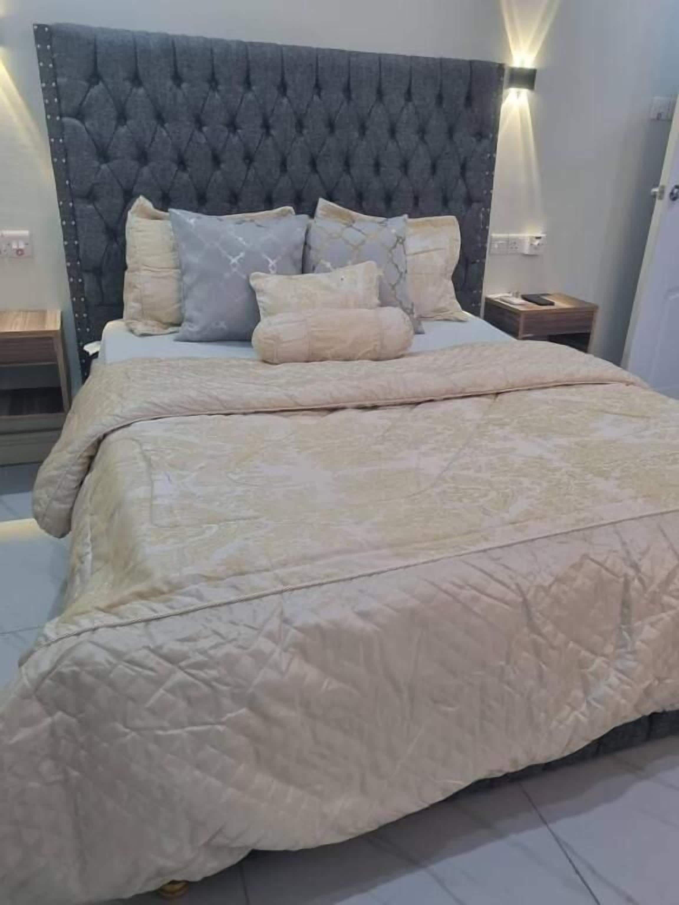Un lit doté d’une tête de lit capitonnée, de plusieurs oreillers et d’une grande couvre-lit beige matelassée.