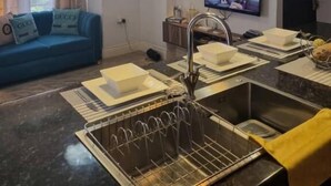 Una sala moderna con un sofá azul, un televisor de pantalla plana, lámparas colgantes, un área de cocina con un lavamanos y un tendedero, y un ventanal con cortinas.
