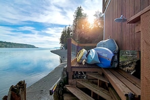 Une terrasse en bois offrant une vue sur un lac paisible et une rive lointaine, un espace abrité avec une douche, ainsi qu’un kayak et un casque sur la terrasse.