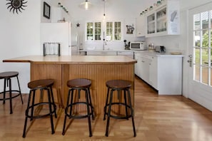 Une cuisine avec un bar en bois, quatre tabourets, des placards blancs, un four à micro-ondes et une fenêtre donnant sur un espace verdoyant.