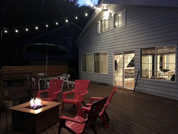 Una terraza con mobiliario exterior y un brasero, una casa con ventanas amplias y luces colgantes.