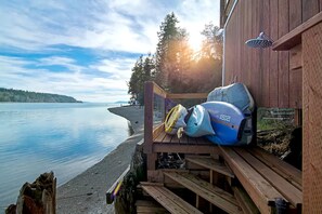 A wooden deck with a view of a calm lake and distant shoreline, a covered area with a shower, and a kayak and helmet on the deck.