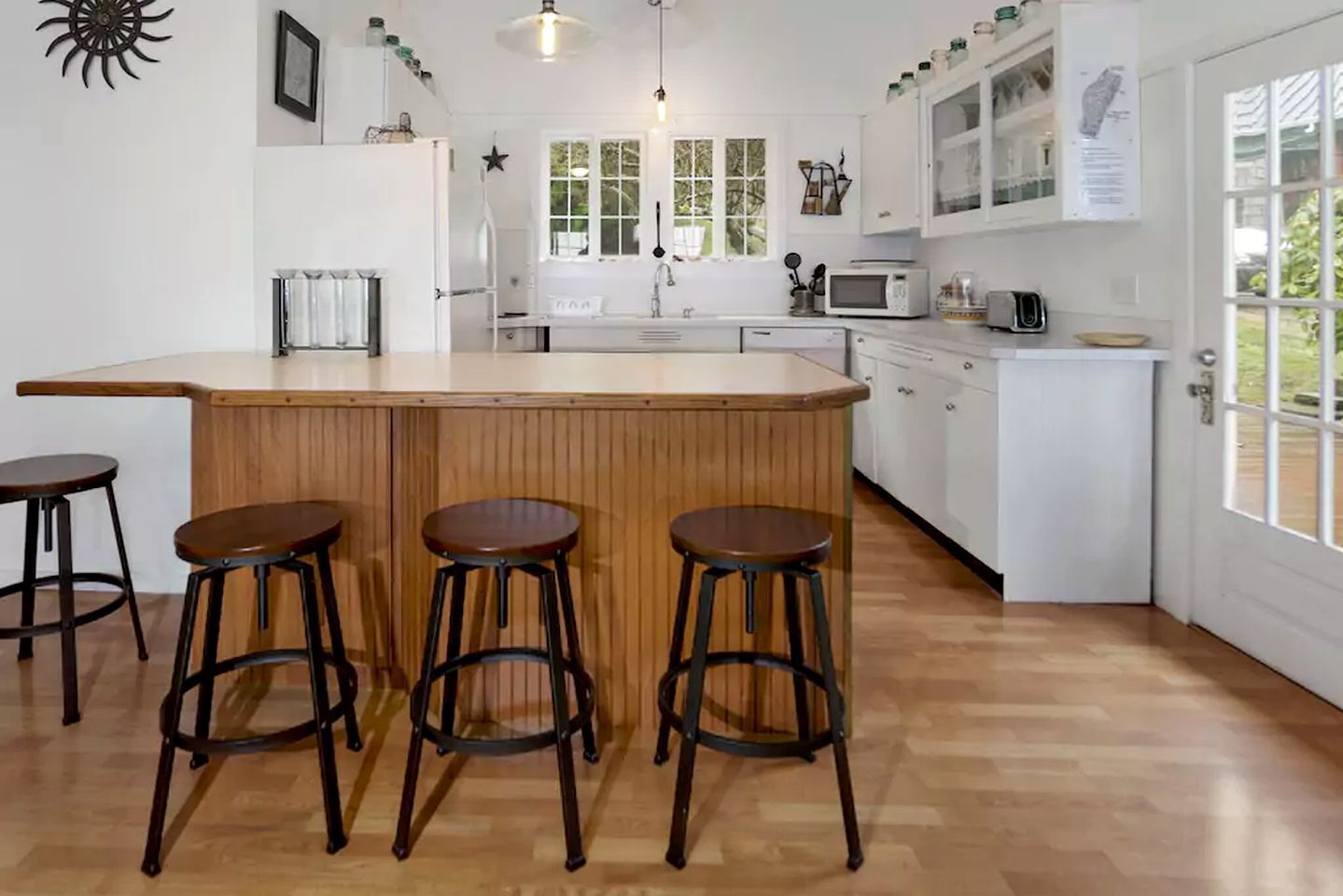 A kitchen with a wooden bar, four stools, white cabinets, a microwave, and a window with greenery outside.