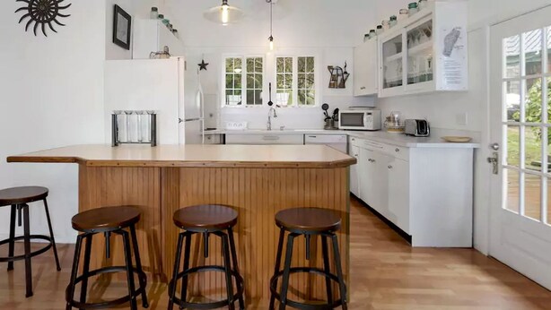 Une cuisine avec un bar en bois, quatre tabourets, des placards blancs, un four à micro-ondes et une fenêtre donnant sur un espace verdoyant.