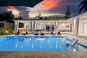 A modern outdoor pool area with lounge chairs, a covered seating area, and a view of the sunset.