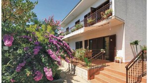 A two-story building with a balcony, a staircase, and a garden with purple flowers.