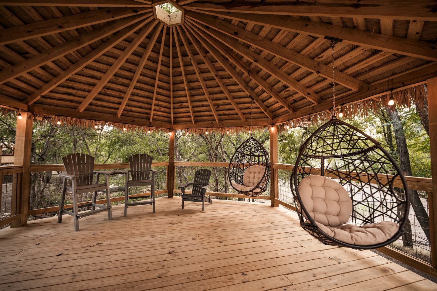 A wooden gazebo with a thatched roof, hanging chairs, and wooden deck.