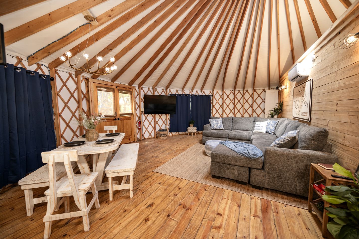 Het interieur van een joert met een houten vloer, een bank, een eettafel met stoelen en een tv aan de wand.