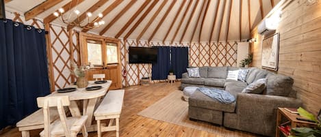 Interior de una yurta con suelo de madera, un sofá, una mesa de comedor con sillas y un televisor colgado en la pared.