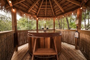 Un quiosco de madera con una mesa redonda y sillas, rodeado de una terraza de madera y una cerca de bambú, con vista a un área boscosa.
