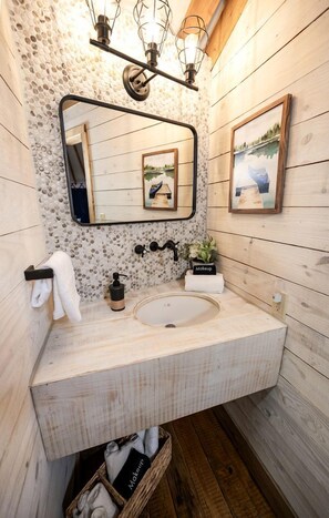 Une salle de bain rustique, avec des murs en bois, comprenant un lavabo rond, un miroir, un porte-serviettes, un distributeur de savon et une petite plante.
