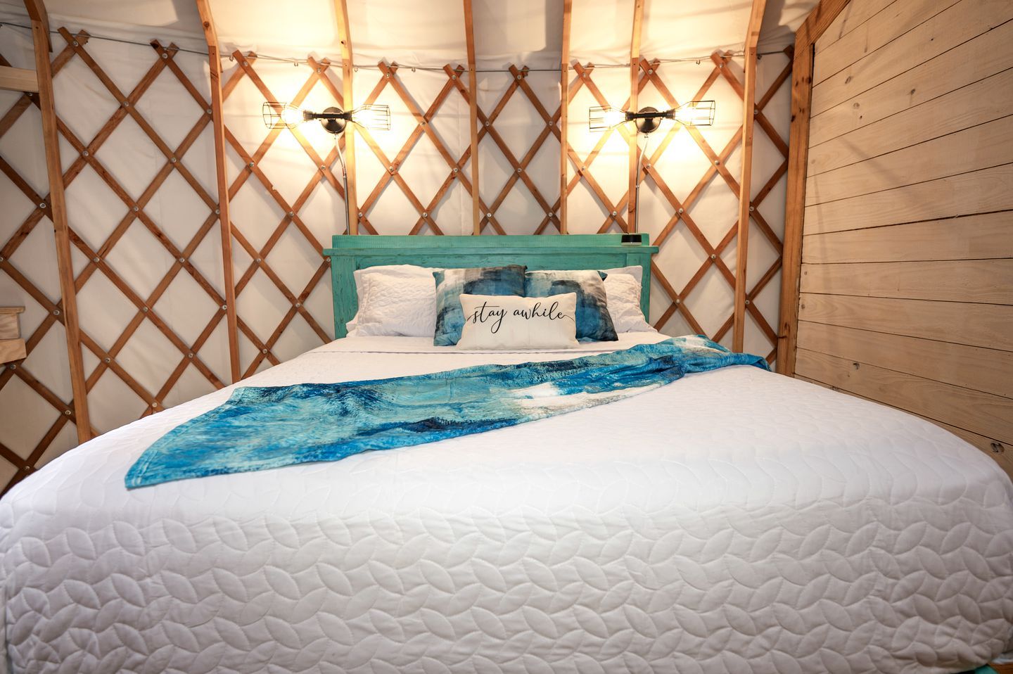 Una cama con colcha blanca, un plaid azul abstracto y un cojín con el texto 'stay awhile'.