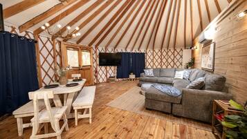A yurt interior with a wooden floor, a sofa, a dining table with chairs, and a TV mounted on the wall.