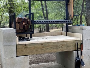Un barbecue extérieur en béton, agrémenté d’un banc en bois, le tout entouré d’arbres.