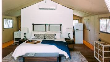 A tented accommodation room with a bed, bedside tables, a lamp, a refrigerator, and a window.