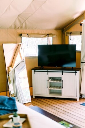 Una habitación con carpa, un televisor de pantalla plana sobre un soporte de madera, un armario blanco con estantes y una ventana con cortinas.