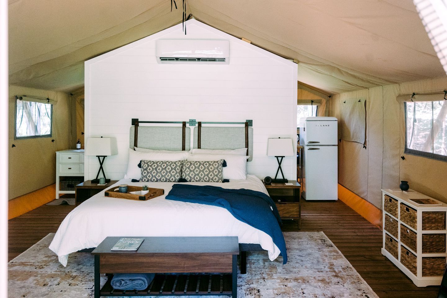 Une chambre aménagée sous une tente, comprenant un lit, des tables de chevet, une lampe, un réfrigérateur et une fenêtre.