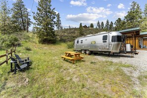 A recreational vehicle parked on a gravel area with a picnic table and chairs, surrounded by trees and a wooden structure. - Fully-Equipped Airstream Rental for Glamping in Chelan, Washington (Chelan)