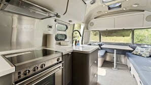 A compact kitchen with a stove, oven, and sink, a small dining area with a bench, and a window providing a view of greenery.