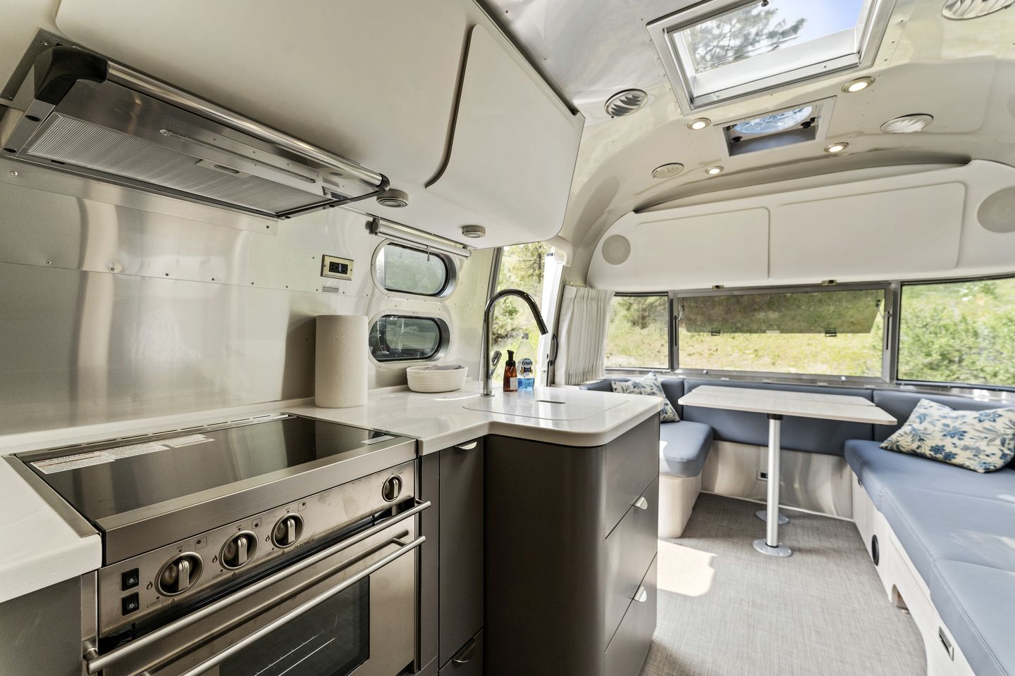 Una cocina compacta con estufa, horno y lavamanos, un pequeño comedor con un banco, y una ventana que ofrece vistas a zonas verdes.