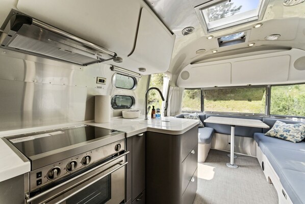 A compact kitchen with a stove, oven, and sink, a small dining area with a bench, and a window providing a view of greenery. - Fully-Equipped Airstream Rental for Glamping in Chelan, Washington (Chelan)