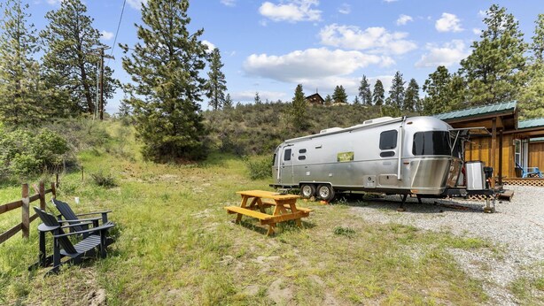 A recreational vehicle parked on a gravel area with a picnic table and chairs, surrounded by trees and a wooden structure.