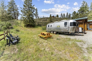 A recreational vehicle parked on a gravel area with a picnic table and chairs, surrounded by trees and a wooden structure. - Fully-Equipped Airstream Rental for Glamping in Chelan, Washington (Chelan)