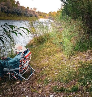 A person seated by a riverbank, wearing a hat and a jacket, with a chair and a striped blanket nearby.