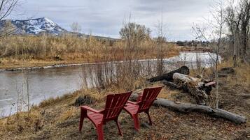 Two red Adirondack chairs by a river with a mountain in the background.