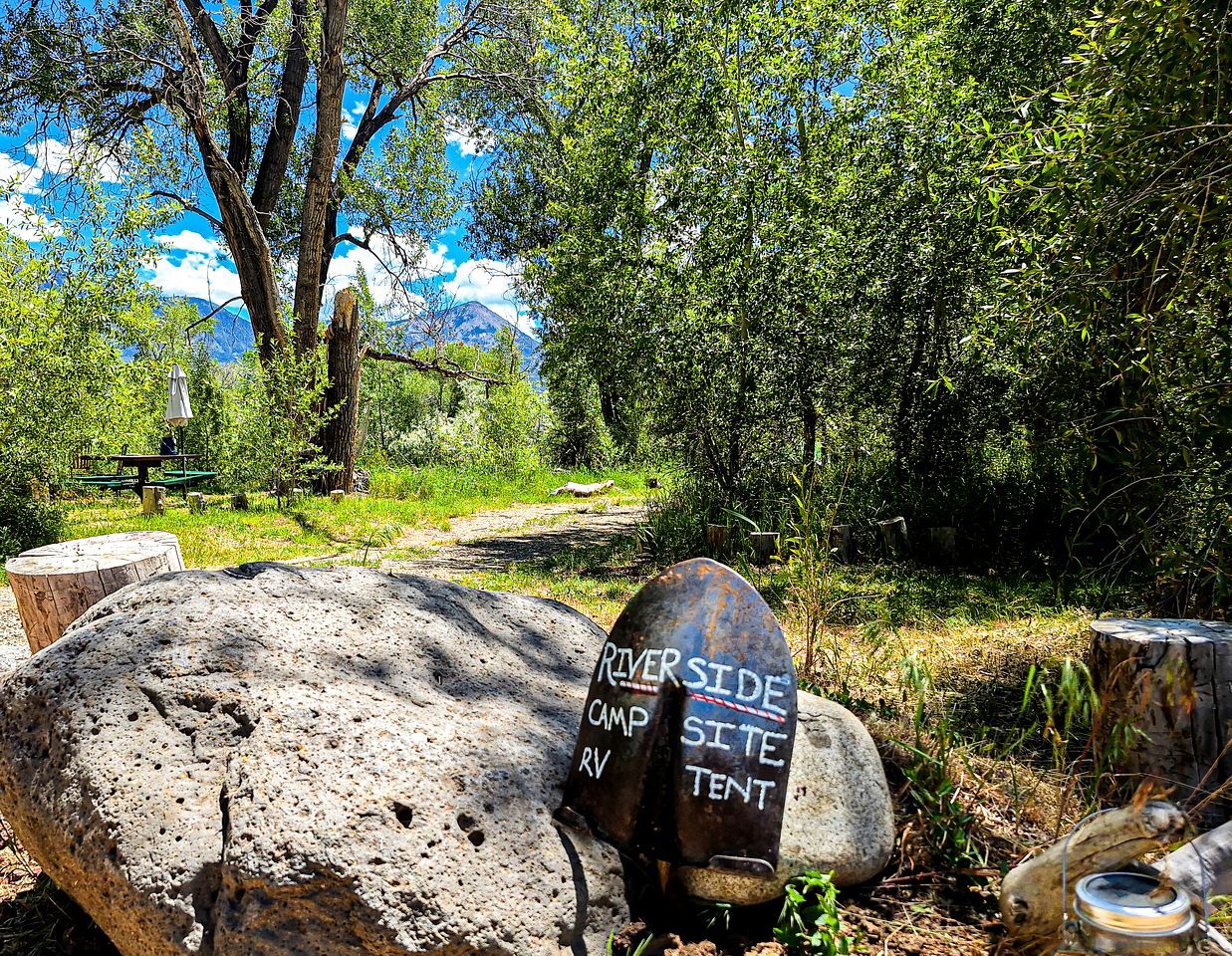 A campsite sign with 'RIVERSIDE CAMP RV TENT' written on it, surrounded by trees and a picnic table.