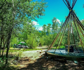 Una estructura tipi tradicional con zona de estar circular y una mesa de comedor, rodeada de árboles y un espacio para picnic.