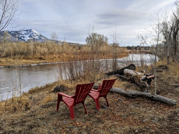 Two red Adirondack chairs by a river with a mountain in the background.